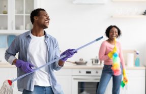 Playful black guy imitating rockstar while house-keeping with his girlfriend, using mop as guitar. Joyful african american couple having fun while cleaning kitchen together, copy space