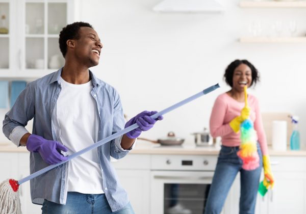 Playful black guy imitating rockstar while house-keeping with his girlfriend, using mop as guitar. Joyful african american couple having fun while cleaning kitchen together, copy space