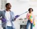 Playful black guy imitating rockstar while house-keeping with his girlfriend, using mop as guitar. Joyful african american couple having fun while cleaning kitchen together, copy space